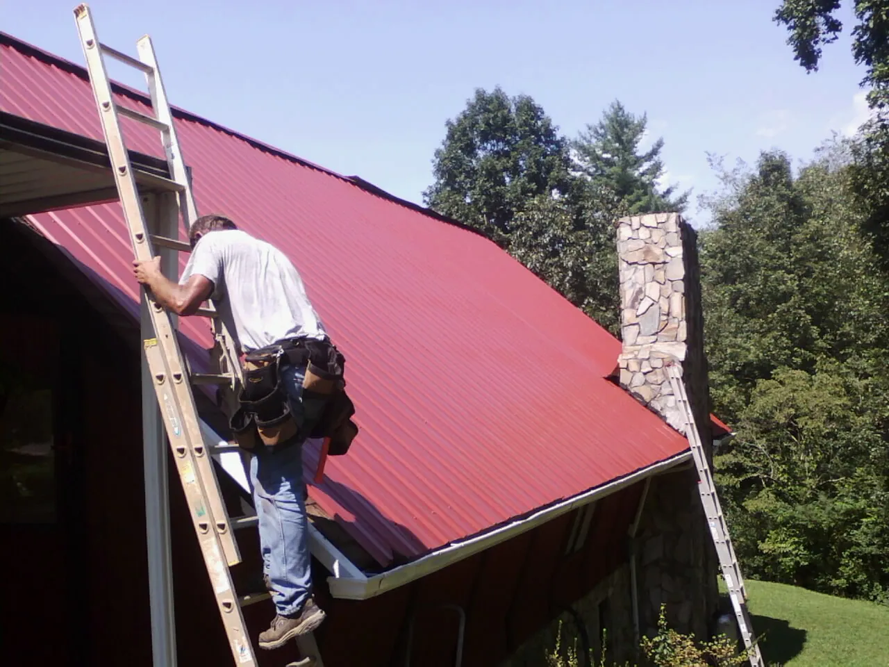 Roofer on a ladder beside a red metal roof and stone chimney — Soffit & Fascia Repair by Roofing Brothers in Pylesville